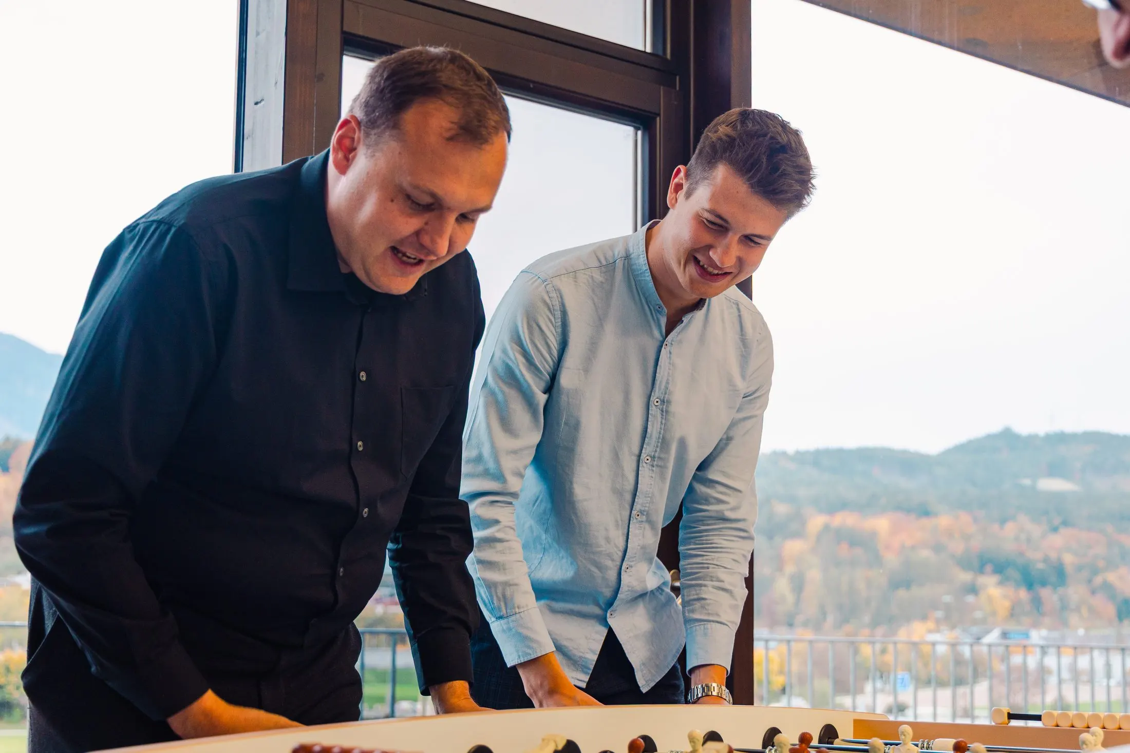 Zwei Kaino-Mitarbeiter spielen Tischfußball vor einem Fenster mit Bergblick